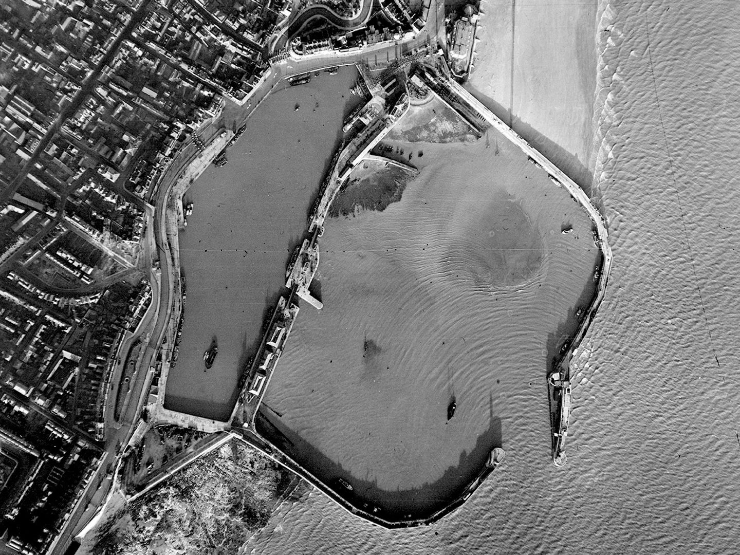 A historic black and white vertical aerial photo of a small harbour. The walls of the harbour reach to the right from the land into the sea, almost meeting and leaving only a narrow entrance for boats. A town is visible on the shore to the left.