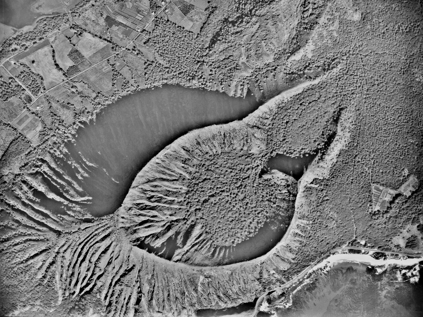 A black and white aerial photo of a heavily forested landscape. An irregular, teardrop-shaped crater rises from the middle of the forest with heavily fluted cliffs. There are fields and a road in the top left of the image, and the sea bottom right.