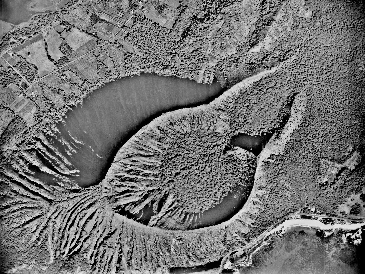 A black and white aerial photo of a heavily forested landscape. An irregular, teardrop-shaped crater rises from the middle of the forest with heavily fluted cliffs. There are fields and a road in the top left of the image, and the sea bottom right.