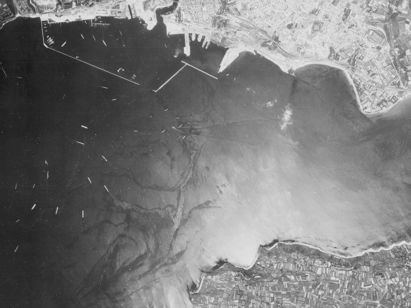 A historic black and white vertical aerial photo of an estuary. Areas of built-up land are visible at the top and bottom edges of the image. Numerous vessels are visible on the water.