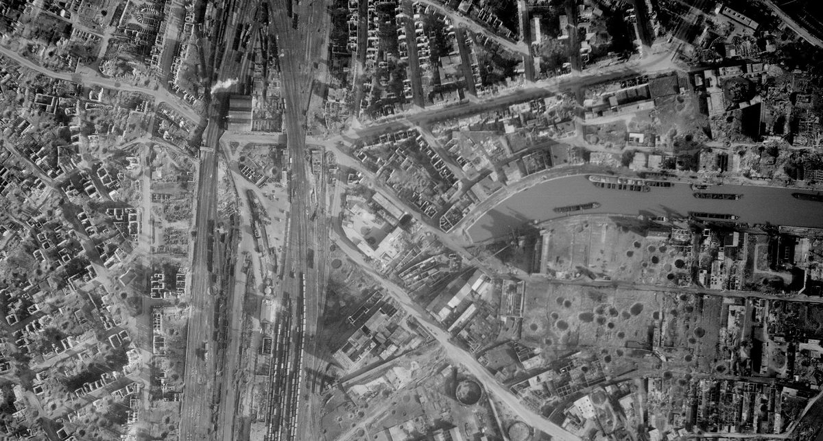 A historic black and white vertical aerial photo of a bombed city. Numerous roofless houses and craters are visible. To the left of the image are trains in some railway sidings. To the right are boats on a canal.