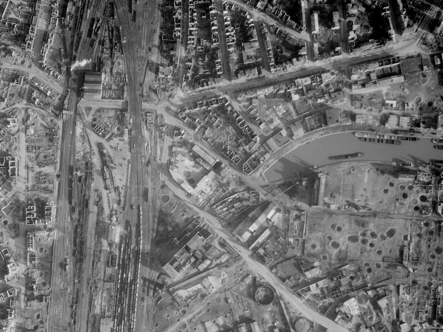 A historic black and white vertical aerial photo of a bombed city. Numerous roofless houses and craters are visible. To the left of the image are trains in some railway sidings. To the right are boats on a canal.