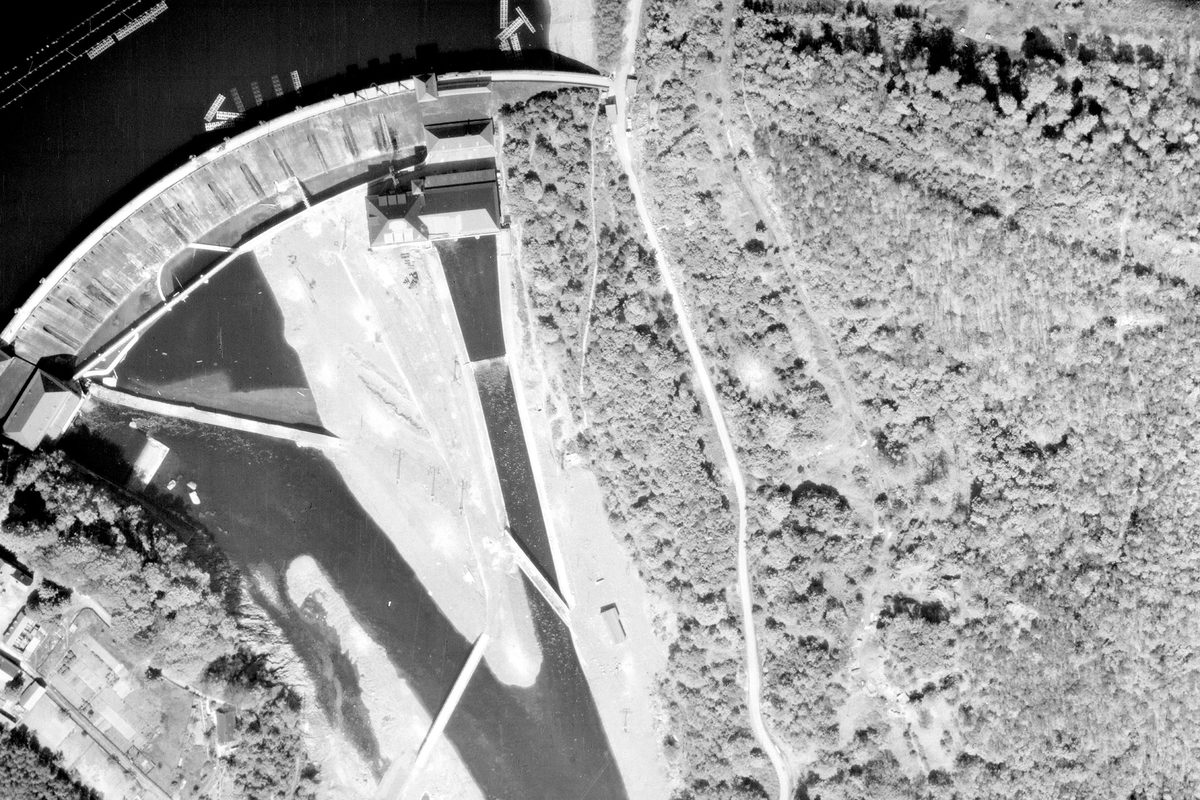 A historic black and white vertical aerial photo of a dam. The reservoir is visible in the top left corner of the image. the banks of the river course below the dam are heavily forested.