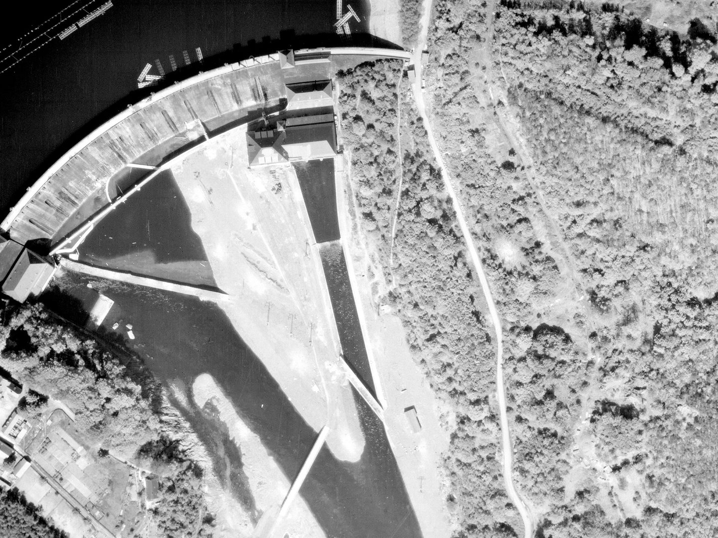 A historic black and white vertical aerial photo of a dam. The reservoir is visible in the top left corner of the image. the banks of the river course below the dam are heavily forested.