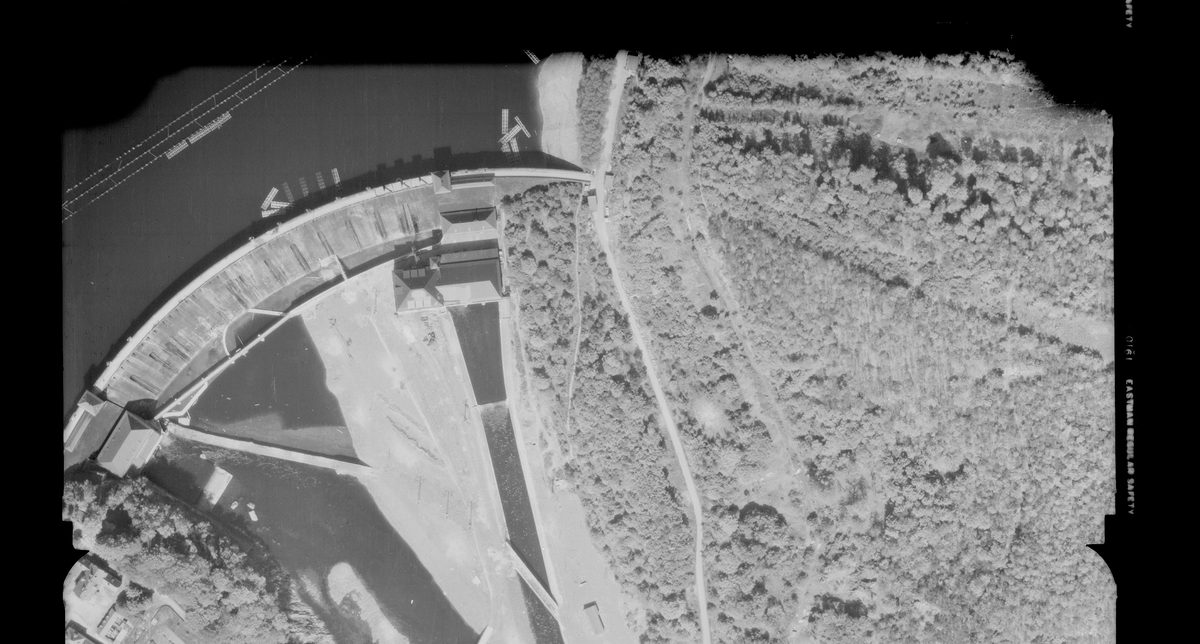 A historic black and white vertical aerial photo of a dam. The dam is to the left of the image, holding back a large body of water. To the right is a large wooded area.