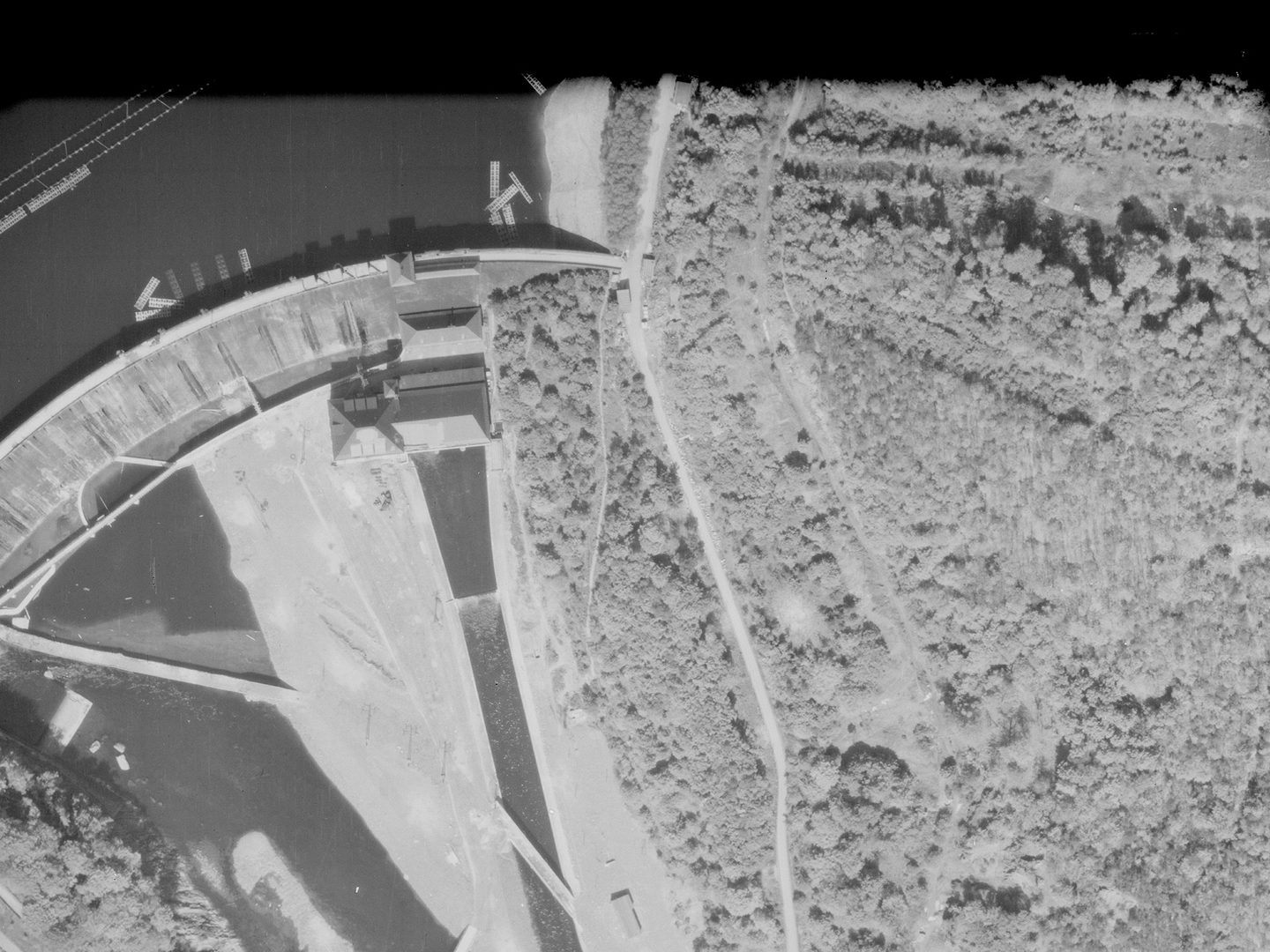 A historic black and white vertical aerial photo of a dam. The dam is to the left of the image, holding back a large body of water. To the right is a large wooded area.