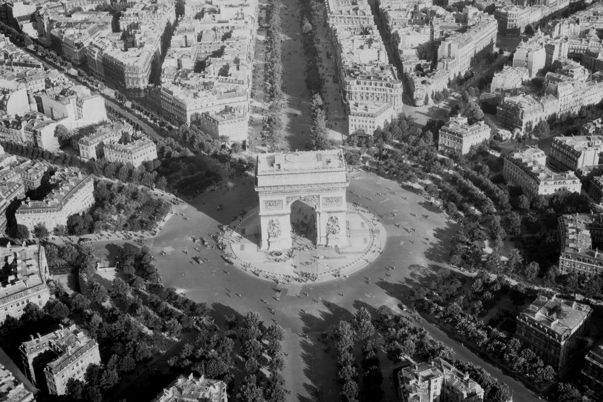A black and white oblique aerial photo of the Arc de Triomphe in Paris. The grand white ceremonial arch stands in the middle of a roundabout. Trees surround the roundabout and handsome white buildings stretch into the distance.