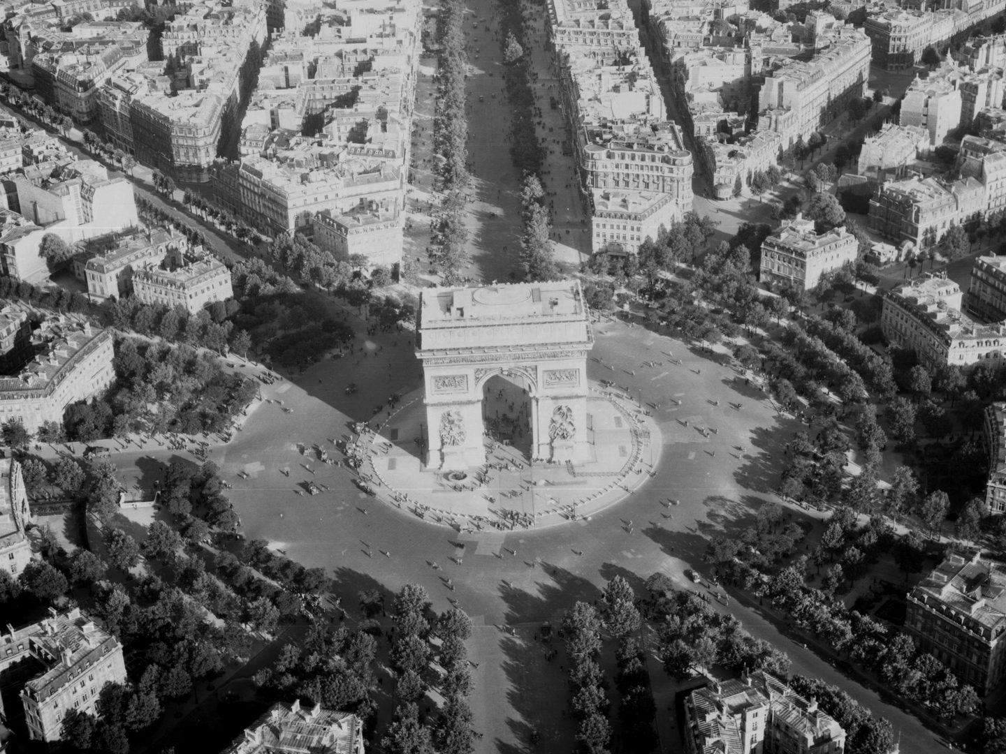 A black and white oblique aerial photo of the Arc de Triomphe in Paris. The grand white ceremonial arch stands in the middle of a roundabout. Trees surround the roundabout and handsome white buildings stretch into the distance.
