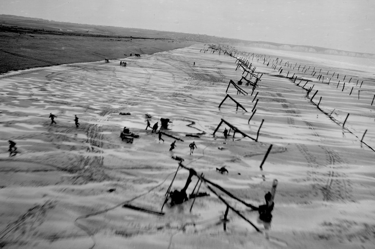A black and white oblique aerial photo of a wide sandy beach. Lines of wooden constructions designed to trap tanks cover the beach, and several figures are visible running and hiding from the approach of the reconnaissance aircraft.