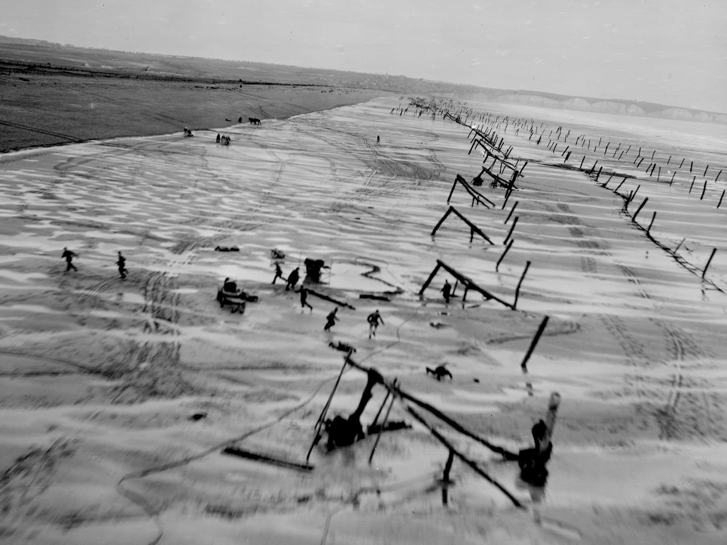 A black and white oblique aerial photo of a wide sandy beach. Lines of wooden constructions designed to trap tanks cover the beach, and several figures are visible running and hiding from the approach of the reconnaissance aircraft.