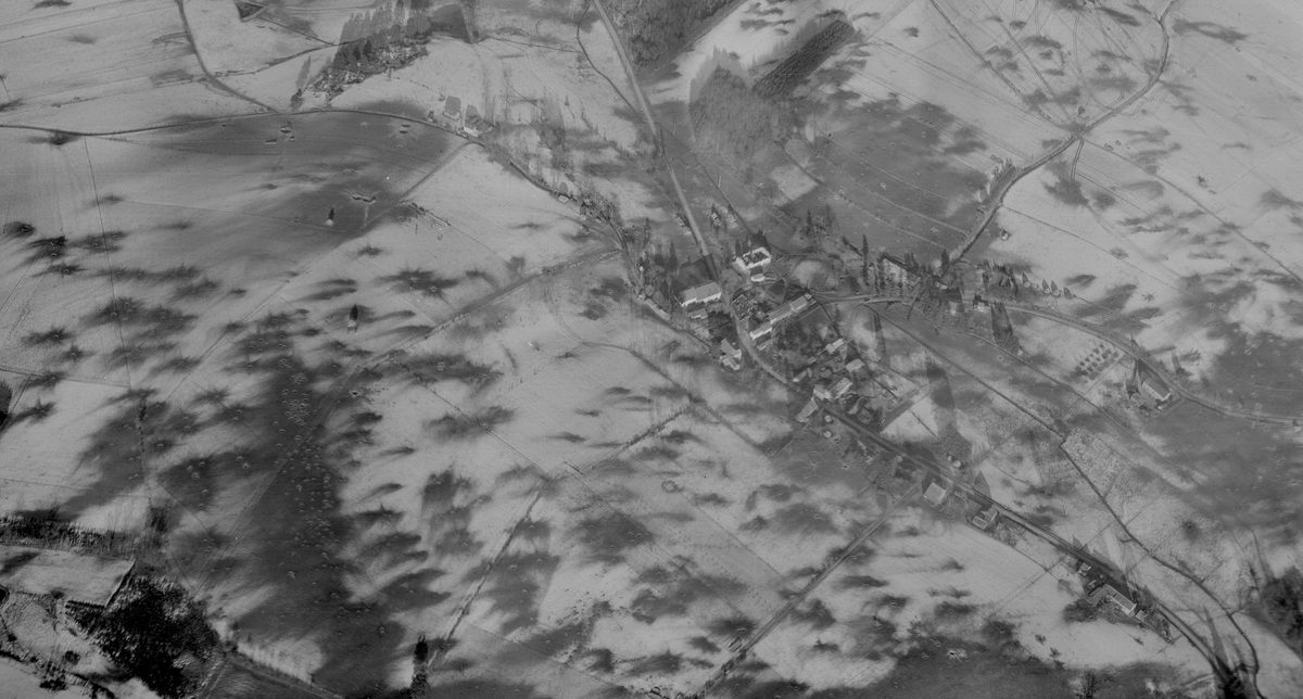 A historic black and white oblique aerial photo of a small village. The village and surrounding fields are covered with snow. Numerous bomb craters mark the snowy fields.
