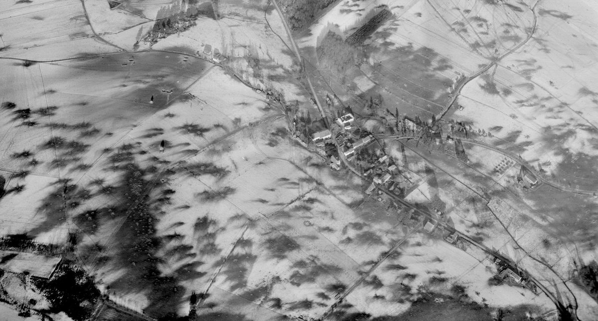A historic black and white oblique aerial photo of a small village. The village and surrounding fields are covered with snow. Numerous bomb craters mark the snowy fields.