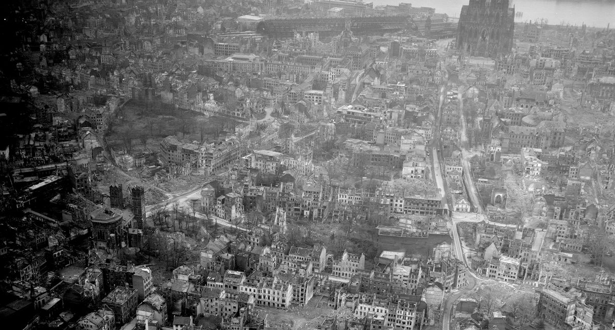 A historic black and white oblique aerial photo of a bombed city. Numerous roofless, bombed houses are visible. In the background is the facade of a large cathedral.