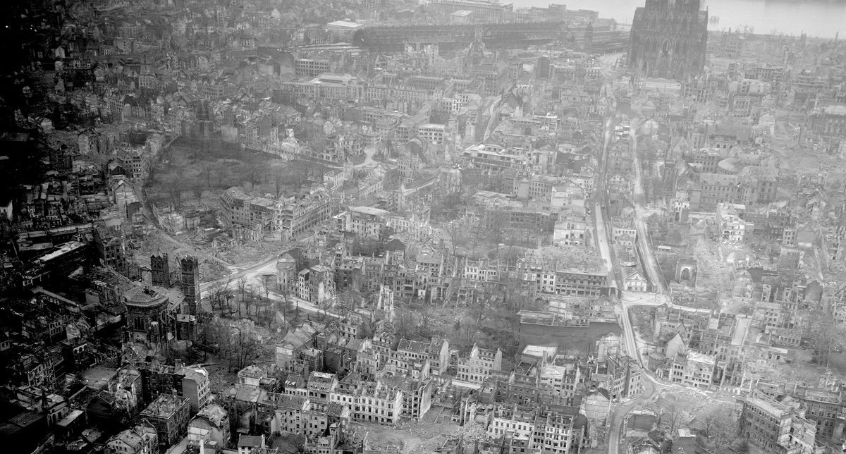 A historic black and white oblique aerial photo of a bombed city. Numerous roofless, bombed houses are visible. In the background is the facade of a large cathedral.