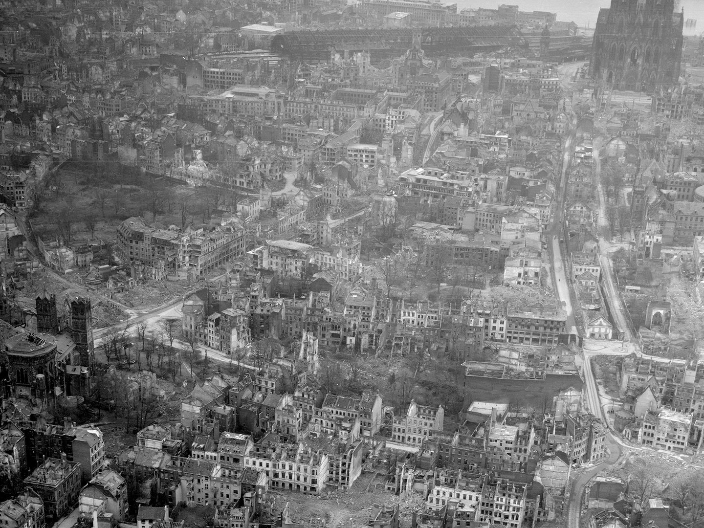 A historic black and white oblique aerial photo of a bombed city. Numerous roofless, bombed houses are visible. In the background is the facade of a large cathedral.
