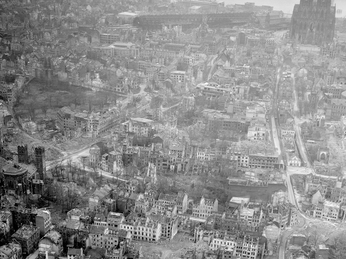A historic black and white oblique aerial photo of a bombed city. Numerous roofless, bombed houses are visible. In the background is the facade of a large cathedral.