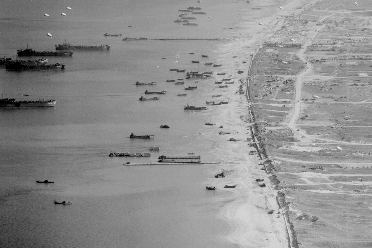 A historic black and white oblique vertical aerial photo of a beach. The sea to the left is covered in vessels of all sizes and descriptions, some offshore and some pulled up on the beach. The land is heavily marked by vehicles and shelling.