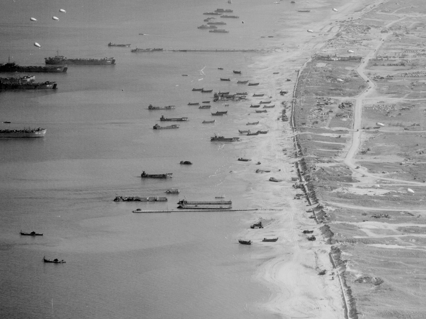 A historic black and white oblique vertical aerial photo of a beach. The sea to the left is covered in vessels of all sizes and descriptions, some offshore and some pulled up on the beach. The land is heavily marked by vehicles and shelling.
