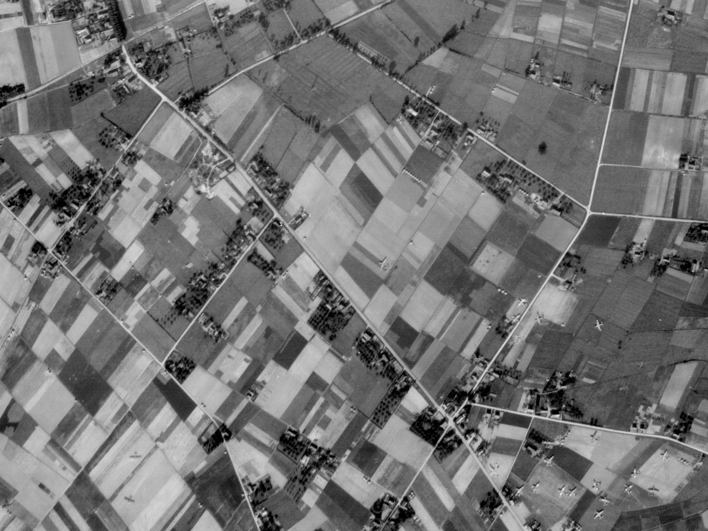A black and white aerial photo of a landscape. Woods run along the left-hand edge of the image. The rest of the image is a patchwork of fields, roads and farms. Scattered across the fields are approximately 30 gliders.
