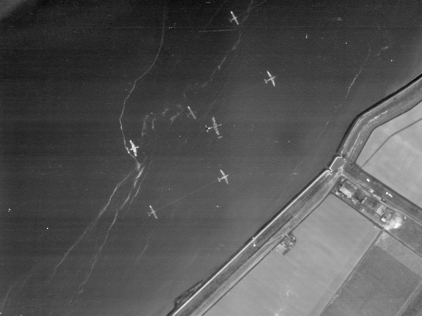 A black and white aerial photo of a coastline, with land in the bottom right corner and the rest of the image filled by sea. Above the sea are 7 aircraft – 3 gliders being towed by 3 planes, and a 4th glider being towed by a plane out of sight.
