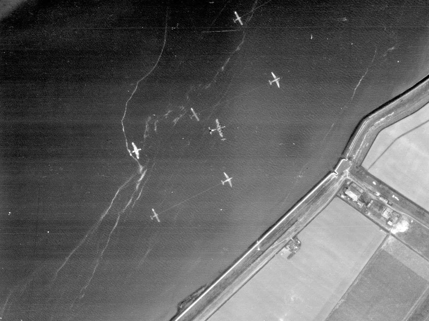 A black and white aerial photo of a coastline, with land in the bottom right corner and the rest of the image filled by sea. Above the sea are 7 aircraft – 3 gliders being towed by 3 planes, and a 4th glider being towed by a plane out of sight.