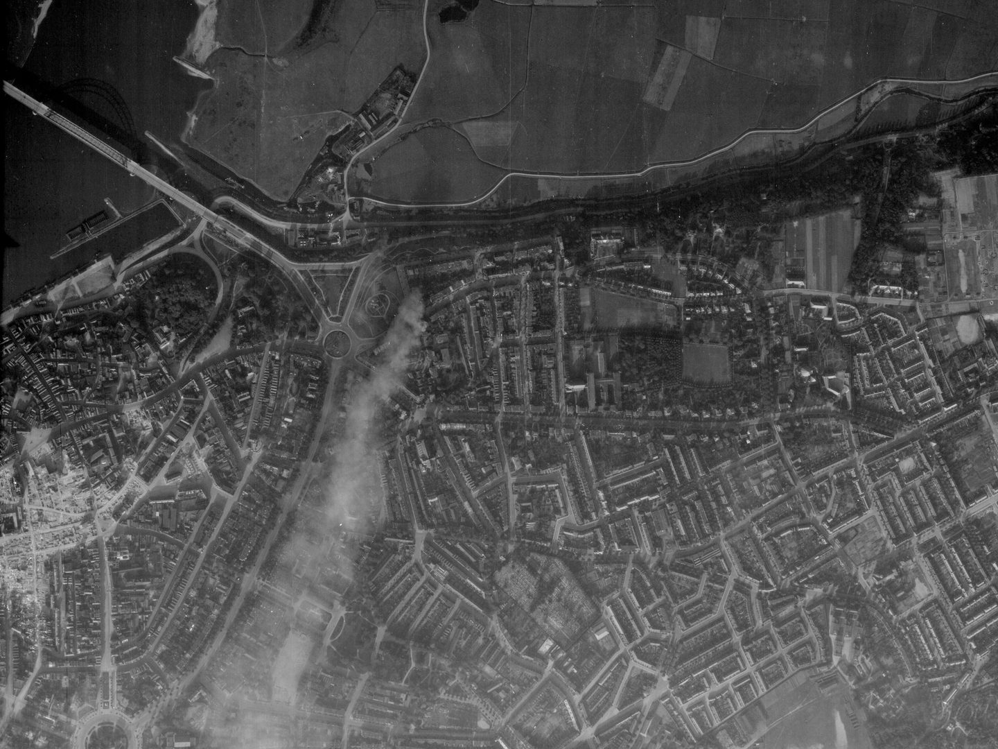 A black and white aerial photo of a town. There are fields along the top edge of the image. In the top right is a wide river crossed by a bridge. The shadow of the bridge is visible on the water. To the left of centre, smoke blows from top to bottom.