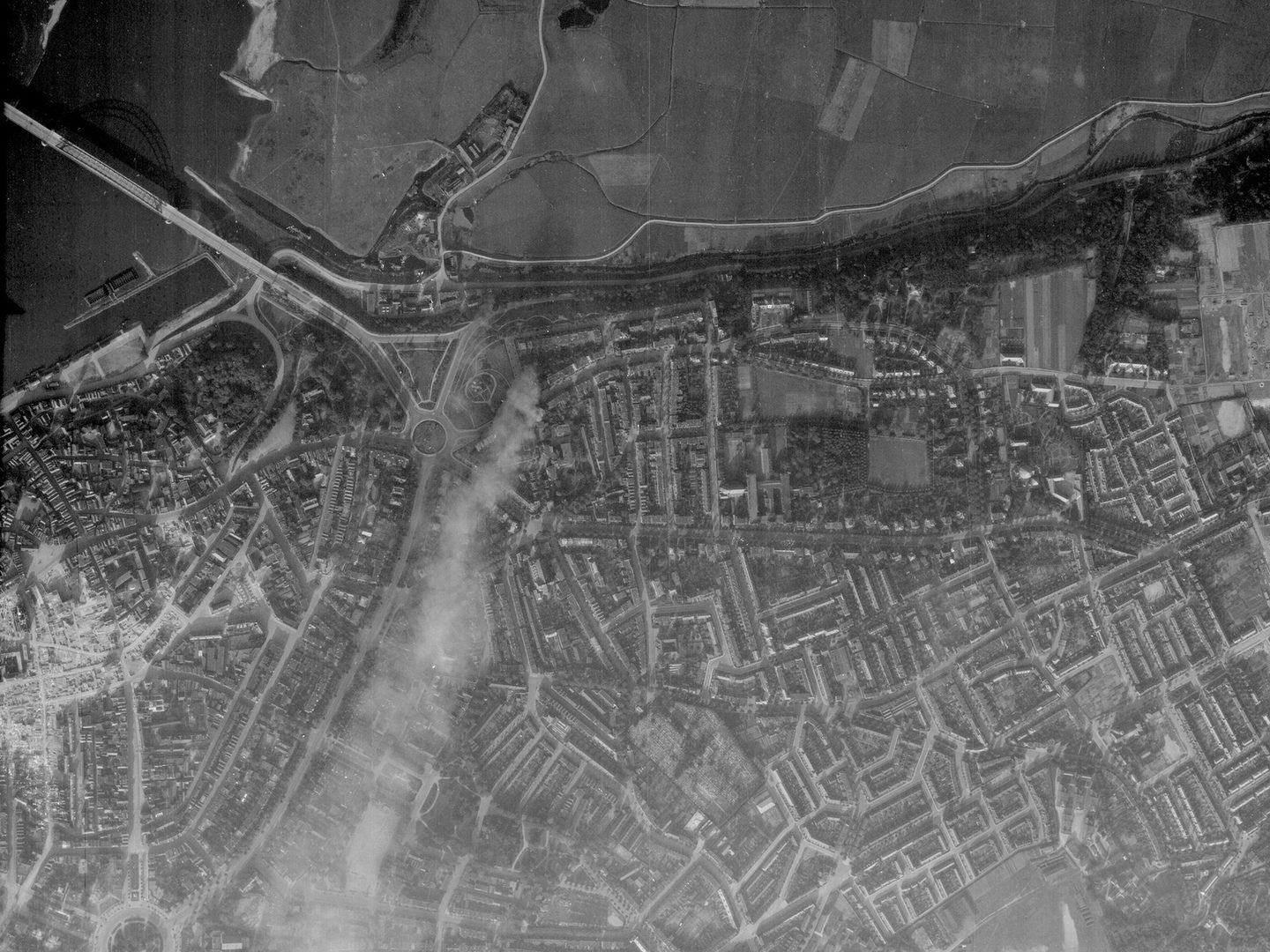 A black and white aerial photo of a town. There are fields along the top edge of the image. In the top right is a wide river crossed by a bridge. The shadow of the bridge is visible on the water. To the left of centre, smoke blows from top to bottom.