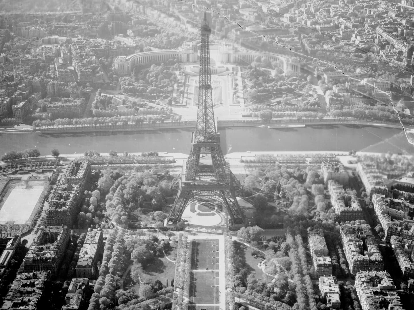 A black and white oblique aerial photo of the Eiffel Tower, surrounded by the streets of Paris, with the River Seine behind.