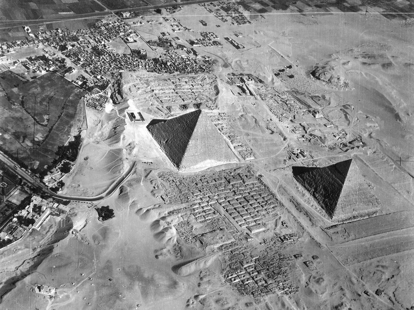 A historic black and white oblique aerial photo of two of the pyramids of Giza, surrounded by sand and other historic structures.