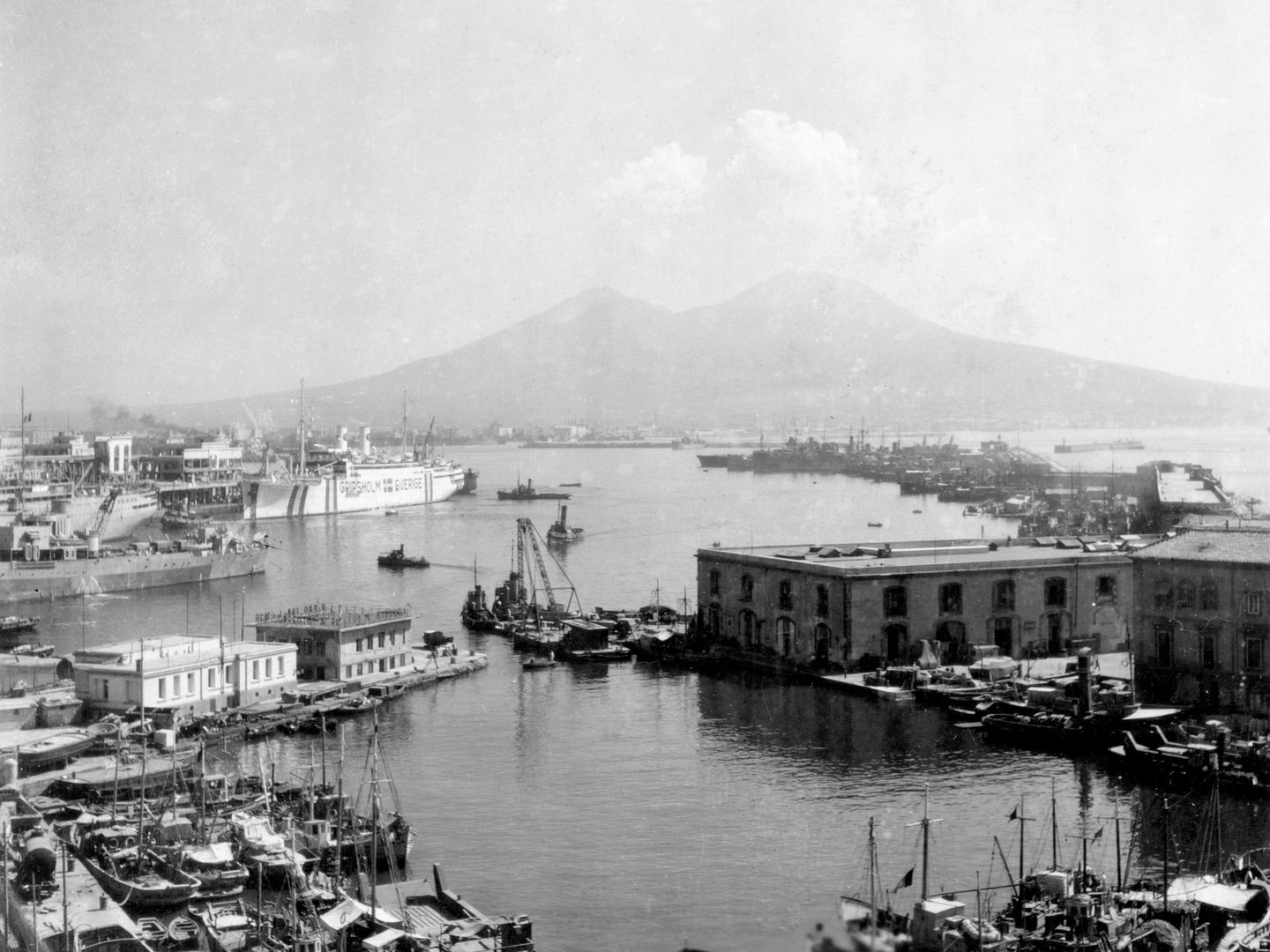 A black and white photo of a port, with numerous small sailing ships in the foreground. A large white liner is visible in the midground, along with several other large ships. In the distance, clouds rise above the double-peak of Mount Vesuvius.