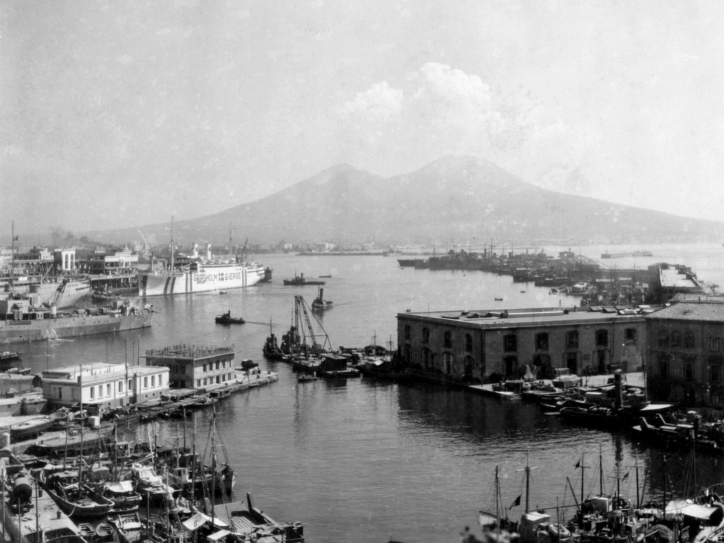 A black and white photo of a port, with numerous small sailing ships in the foreground. A large white liner is visible in the midground, along with several other large ships. In the distance, clouds rise above the double-peak of Mount Vesuvius.