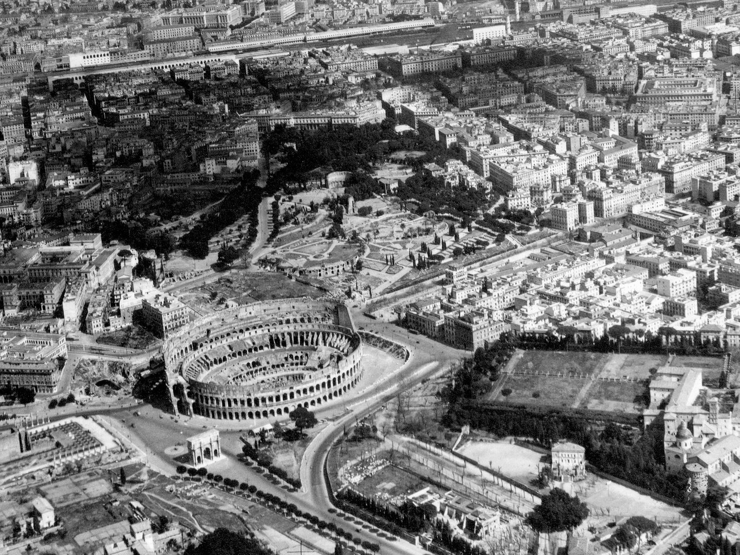 A historic black and white oblique aerial photo of the Coliseum in Rome with Celio Park to the right and Rome Termini Station in the background.