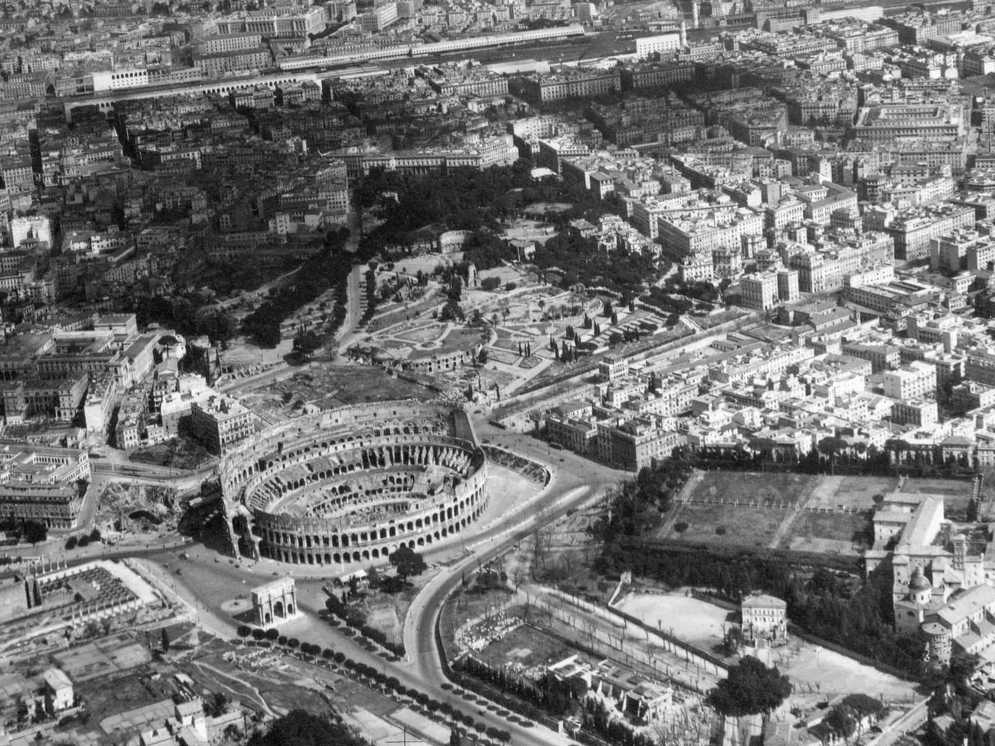 A historic black and white oblique aerial photo of the Coliseum in Rome with Celio Park to the right and Rome Termini Station in the background.