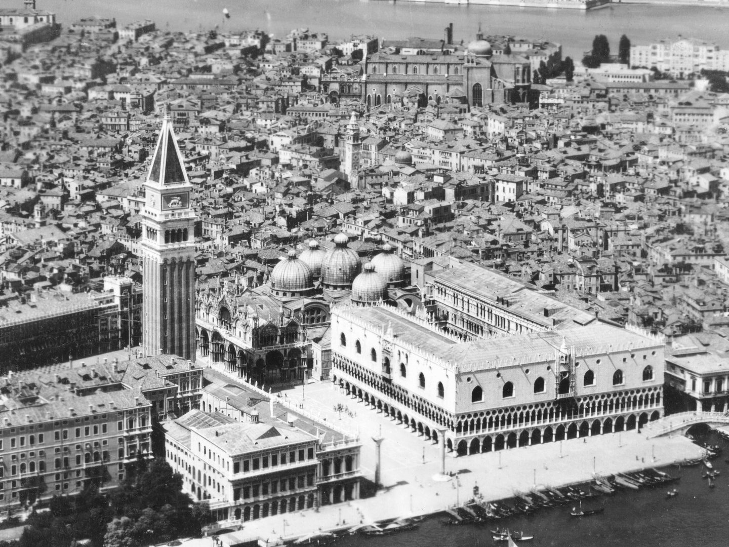 A historic black and white oblique aerial photo of St Mark's Square in Venice, with the Doge's Palace prominent to the right and St Mark's bell tower prominent to the left. The city of Venice is visible behind them.