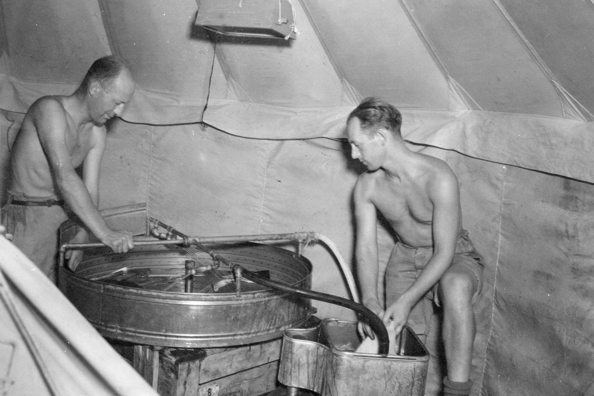 A historic black and white photo of 2 men in shorts and with their shirts off processing photographs using metal tanks in a tent.