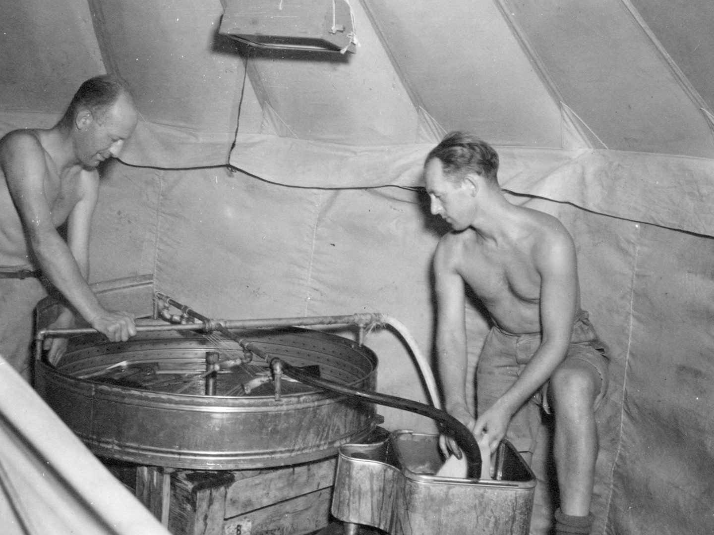 A historic black and white photo of 2 men in shorts and with their shirts off processing photographs using metal tanks in a tent.