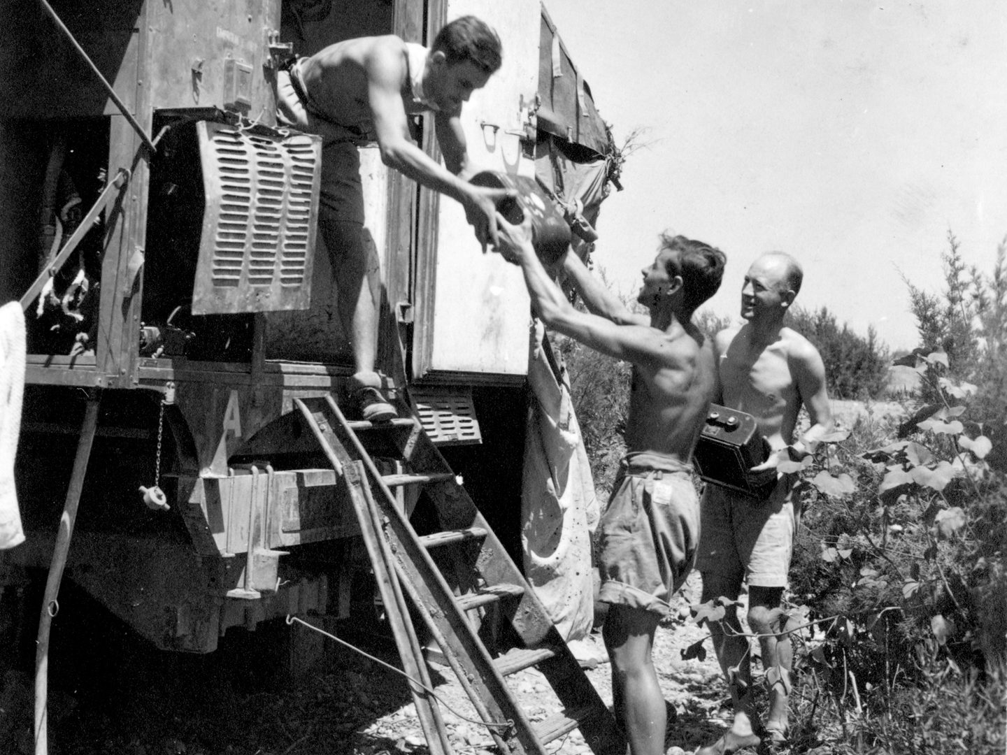 A historic black and white photo of three shirtless men wearing shorts. One man is handing a piece of photographic equipment to another from the doorway of a railway wagon. The third stands in the background holding another piece of photographic equipment.