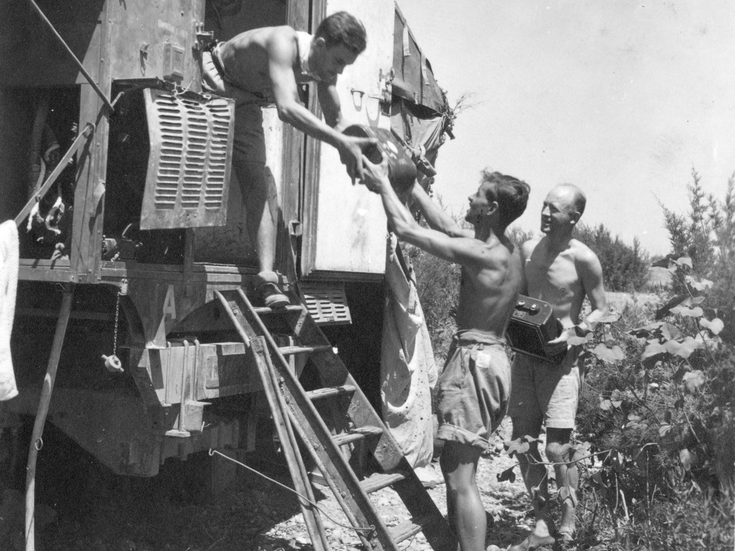 A historic black and white photo of three shirtless men wearing shorts. One man is handing a piece of photographic equipment to another from the doorway of a railway wagon. The third stands in the background holding another piece of photographic equipment.