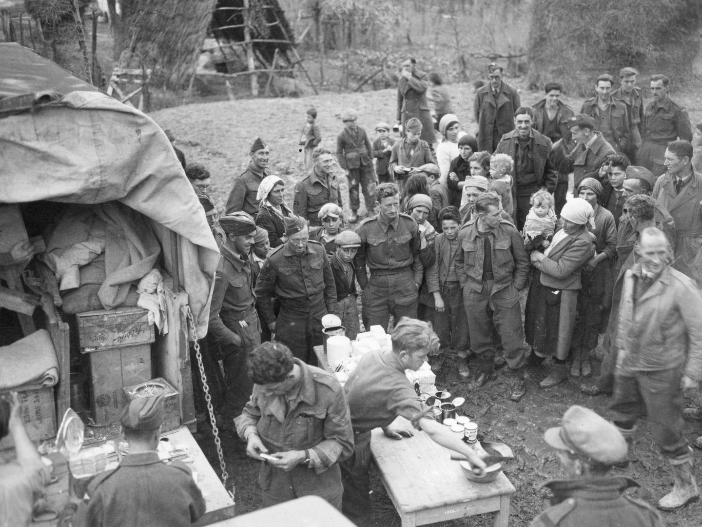 A historic black and white photo of a crowd of people. The crowd is a mix of soldiers in uniform and civilians - adults and children. The focus of the crowd is a table bearing several opened tins. To the left of the image is a truck filled with packing cases.