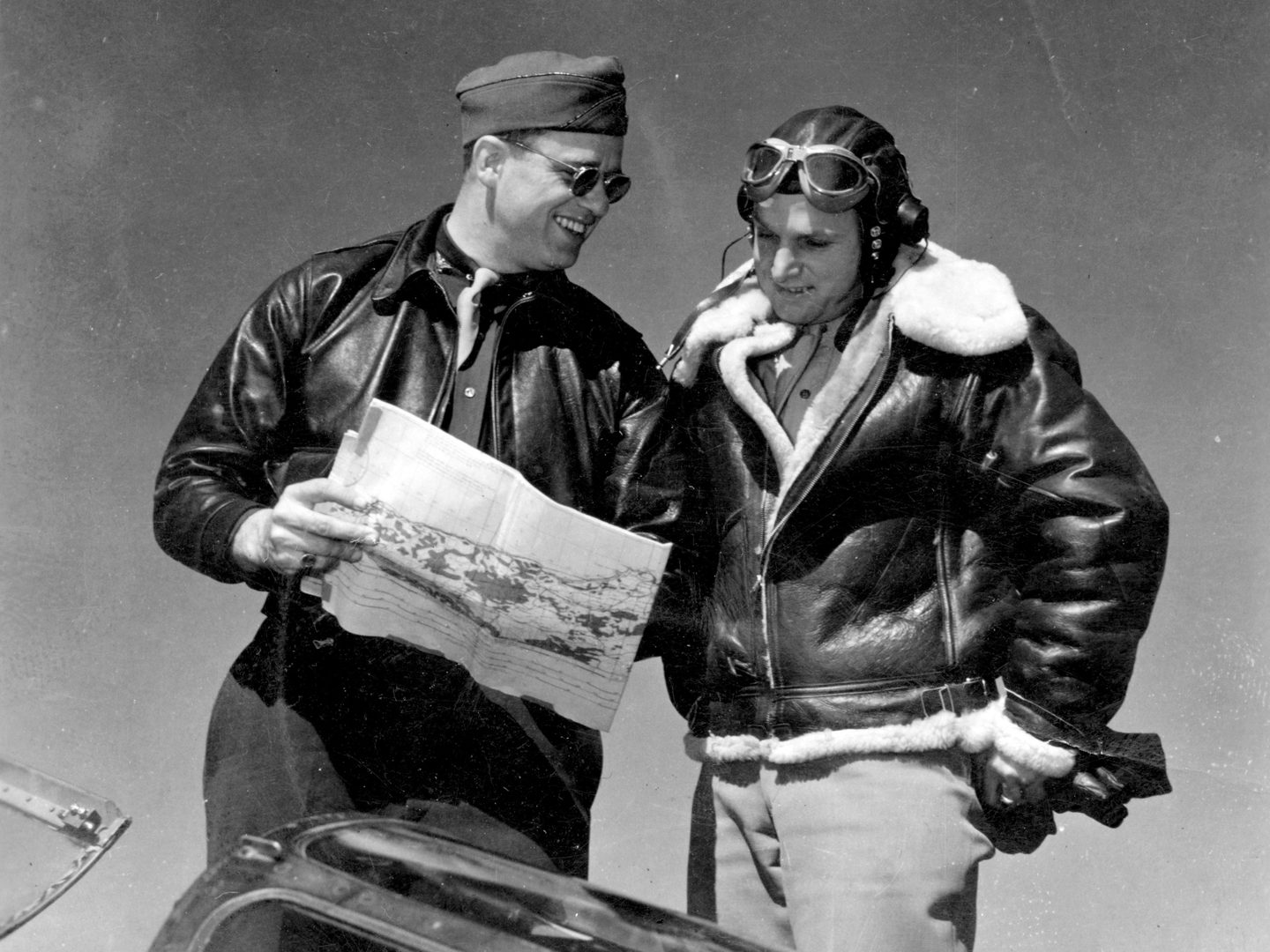 A historic black and white photo of 2 men wearing leather jackets. Roosevelt stands to the left holding a map and wearing sunglasses. The man to the right is wearing a flying cap and goggles, and his jacket is fur lined.