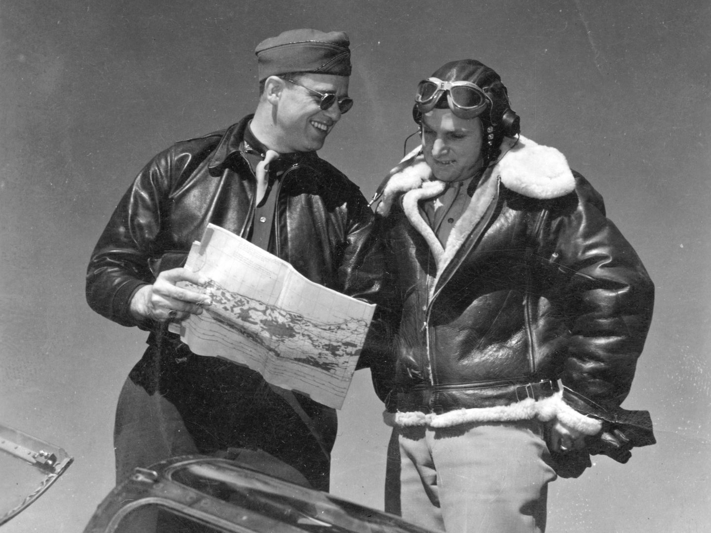 A historic black and white photo of 2 men wearing leather jackets. Roosevelt stands to the left holding a map and wearing sunglasses. The man to the right is wearing a flying cap and goggles, and his jacket is fur lined.
