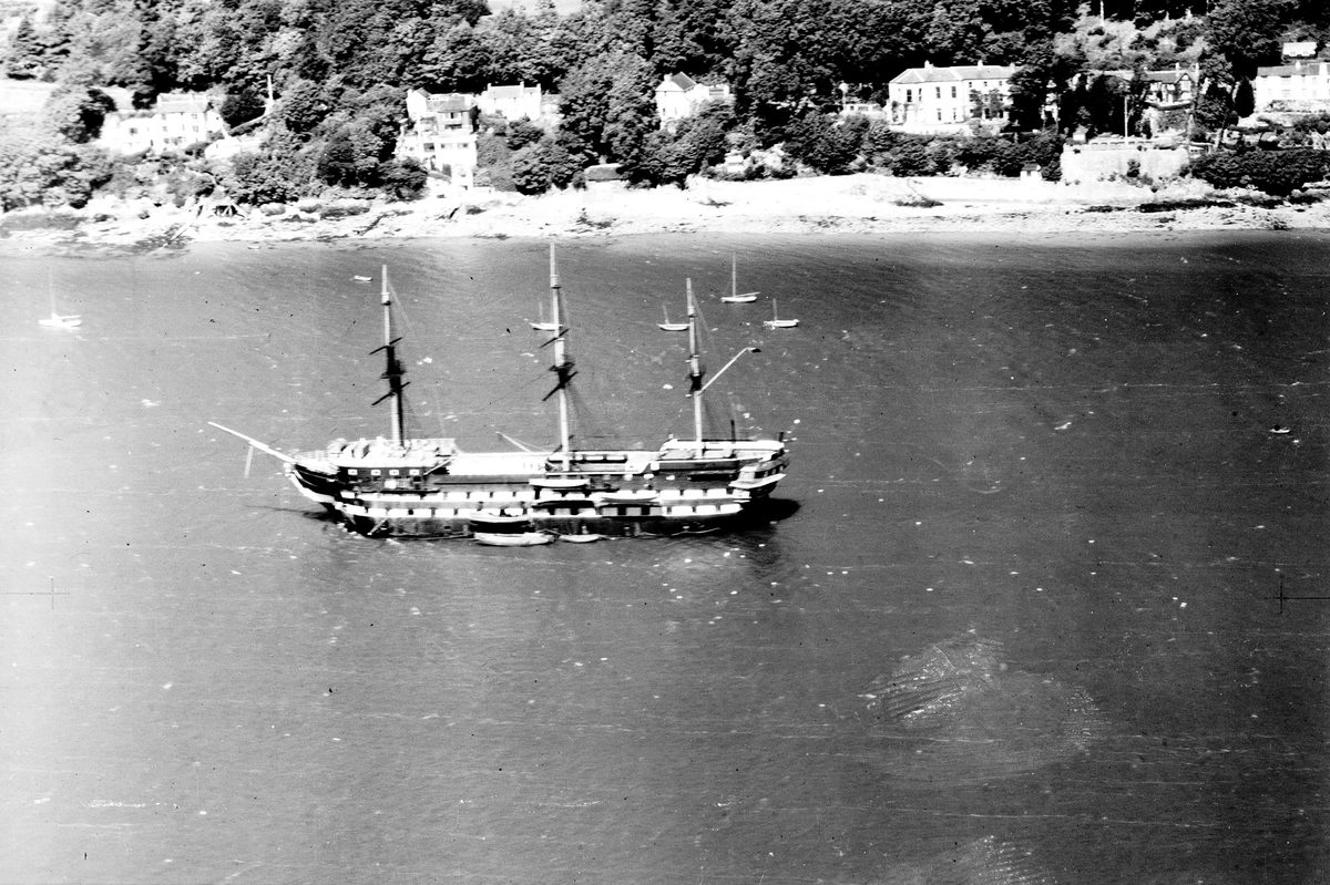 A historic black and white oblique aerial photo of a sailing ship with 3 masts moored off a wooded coast. Several large houses are visible between the trees on the shore.
