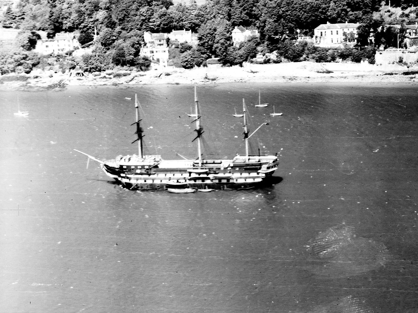 A historic black and white oblique aerial photo of a sailing ship with 3 masts moored off a wooded coast. Several large houses are visible between the trees on the shore.