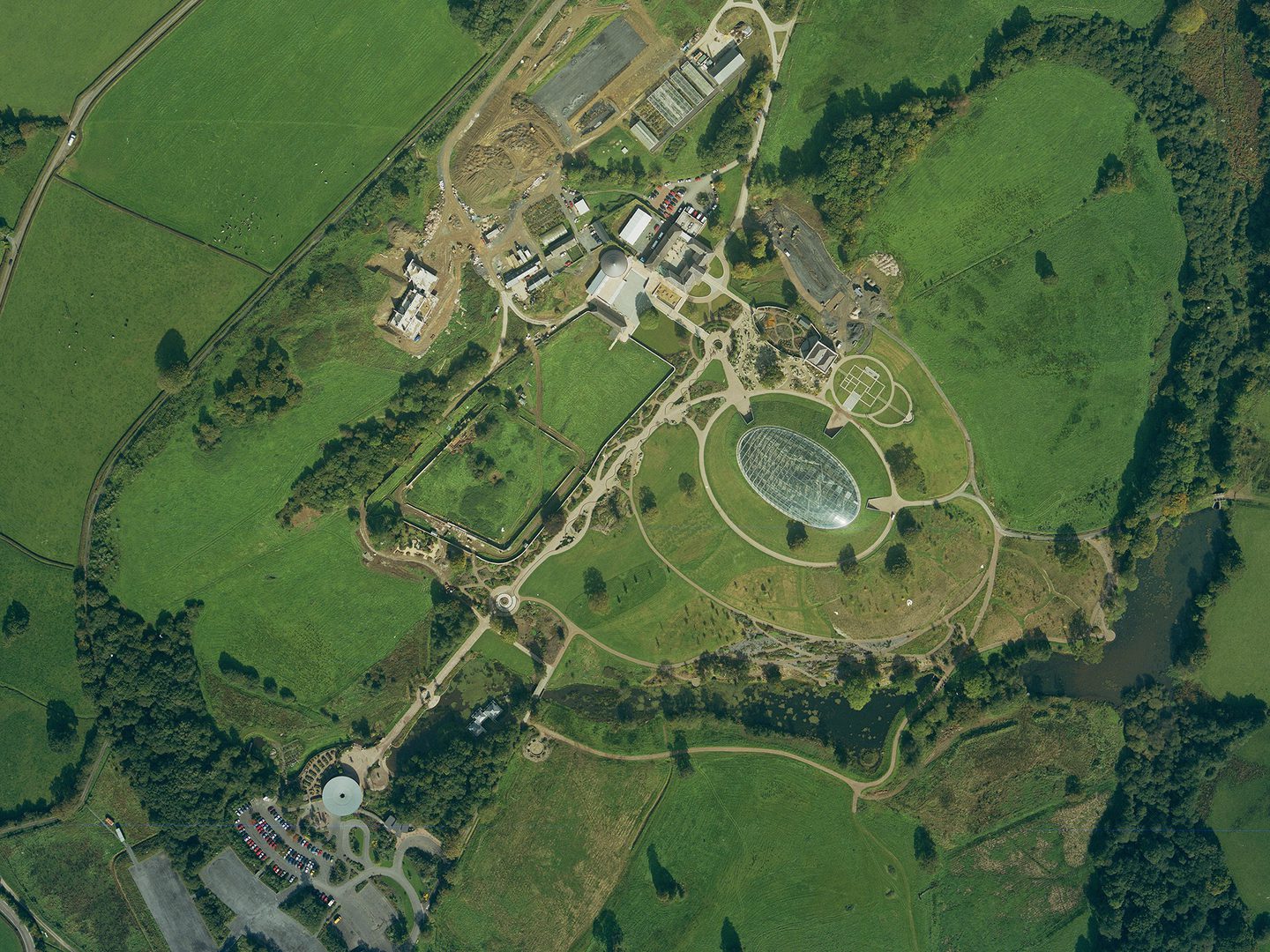 A historic colour vertical aerial photo of an area of countryside with fields and small areas of woodland. In the centre is a very large oval-shaped glass dome. Nearby is a mix of completed buildings and areas of construction.