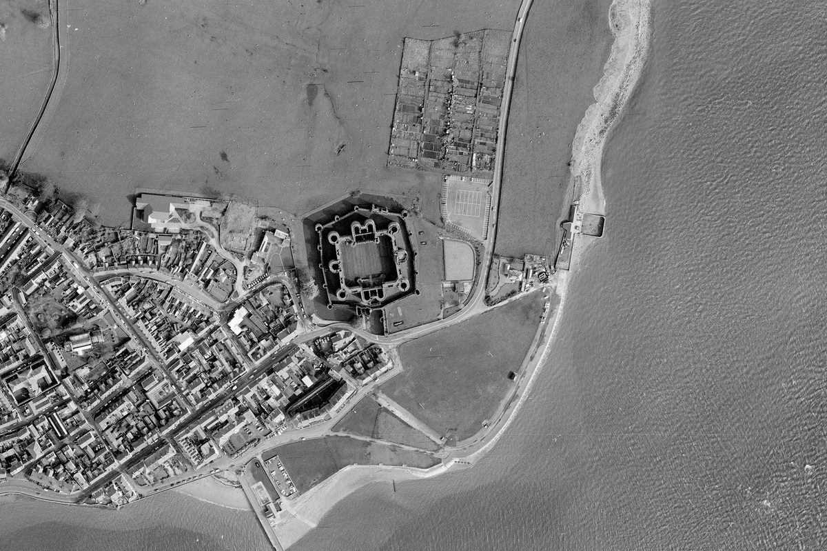 A historic black and white vertical aerial photo of a coastline. The sea runs along the right-hand and bottom edges of the image, with a town visible on the land to the left. In the centre is a large castle, with 2 curtain walls, 2 grand gatehouses and numerous large towers.