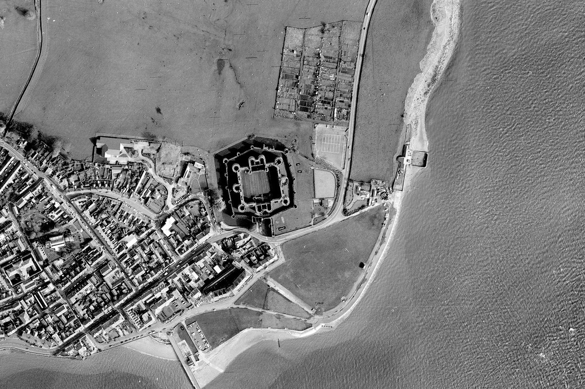 A historic black and white vertical aerial photo of a coastline. The sea runs along the right-hand and bottom edges of the image, with a town visible on the land to the left. In the centre is a large castle, with 2 curtain walls, 2 grand gatehouses and numerous large towers.