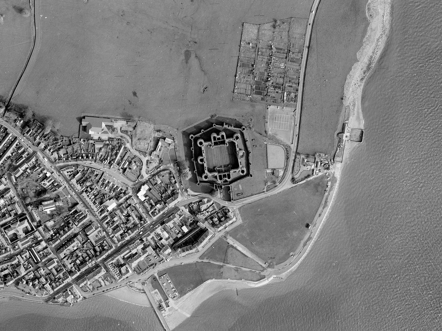 A historic black and white vertical aerial photo of a coastline. The sea runs along the right-hand and bottom edges of the image, with a town visible on the land to the left. In the centre is a large castle, with 2 curtain walls, 2 grand gatehouses and numerous large towers.