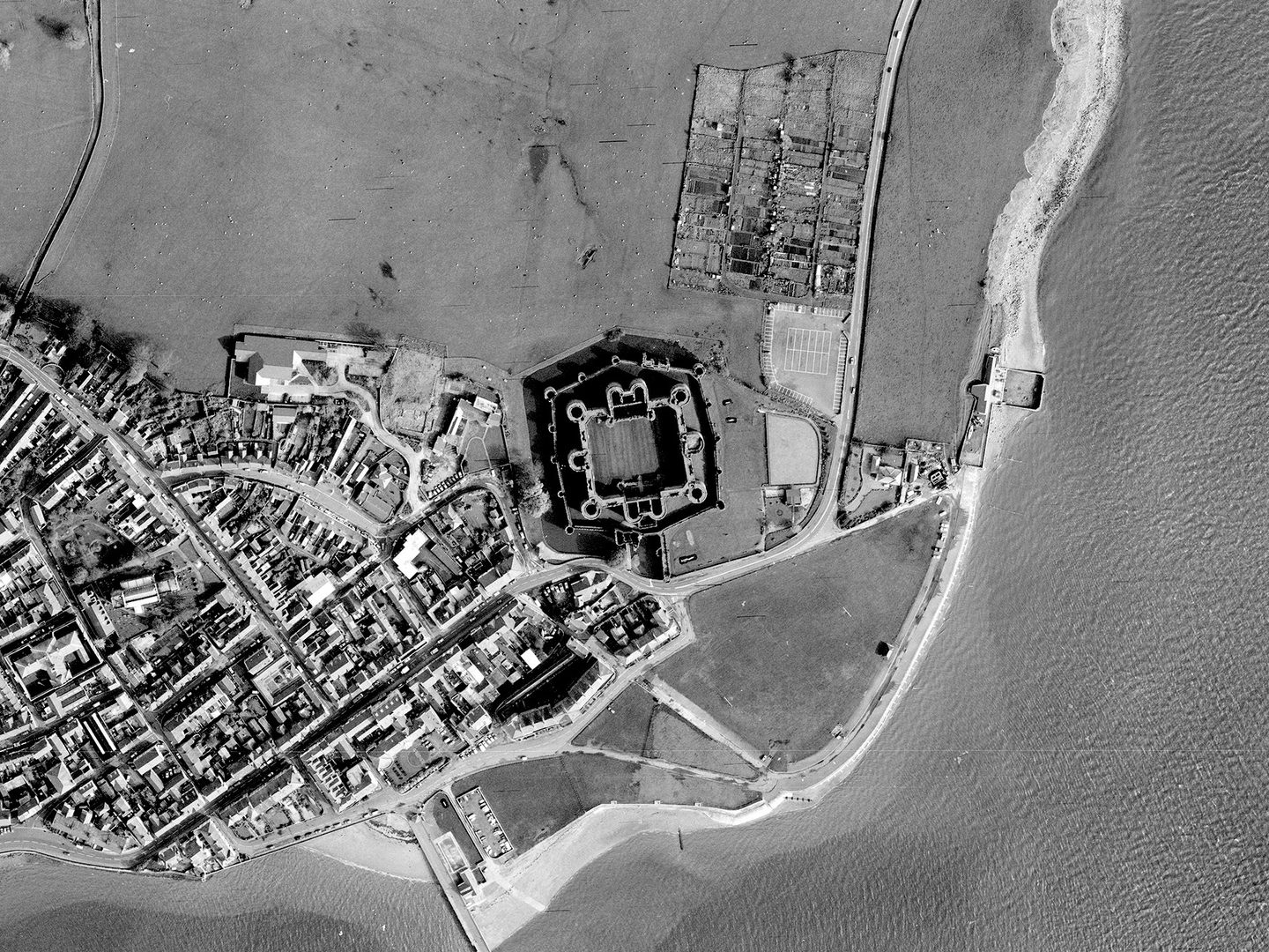 A historic black and white vertical aerial photo of a coastline. The sea runs along the right-hand and bottom edges of the image, with a town visible on the land to the left. In the centre is a large castle, with 2 curtain walls, 2 grand gatehouses and numerous large towers.