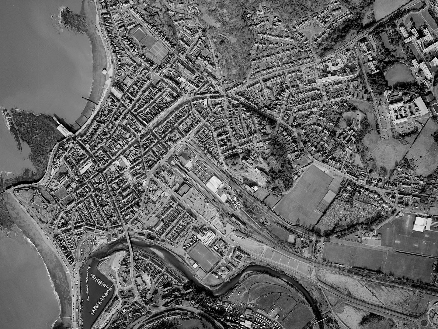 A historic black and white vertical aerial photo of the town of Aberystwyth, with the sea to the left and the town stretching through fields to the right. A river runs along the bottom of the image.