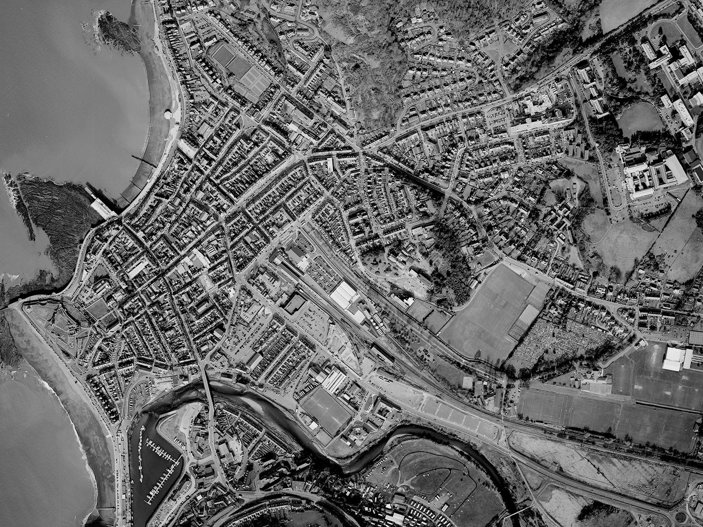 A historic black and white vertical aerial photo of the town of Aberystwyth, with the sea to the left and the town stretching through fields to the right. A river runs along the bottom of the image.